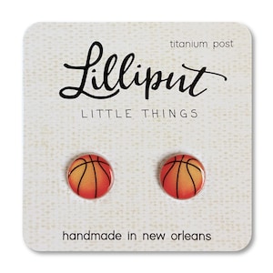 May include: A pair of earrings featuring a basketball design. The earrings are orange and black with a white background. The earrings are handmade in New Orleans and have titanium posts. The card reads "Lilliput Little Things".