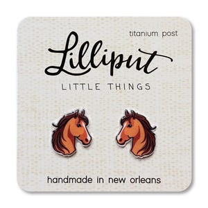 May include: Horse head stud earrings on a white card. The earrings are brown and orange with a titanium post. The card has the text "Lilliput Little Things" and "handmade in new orleans".