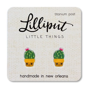 May include: A pair of cactus earrings with a pink flower on top. The earrings are handmade in New Orleans and have titanium posts. The card says "Lilliput Little Things".