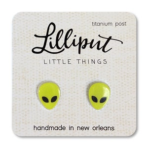 May include: A pair of yellow alien head stud earrings with black eyes. The earrings are on a white card with the text "Lilliput Little Things" and "handmade in new orleans".