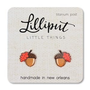 May include: A pair of acorn stud earrings with a red and brown leaf detail. The earrings are handmade in New Orleans and feature titanium posts. The card reads "Lilliput Little Things".