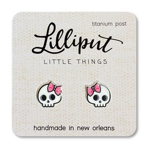 May include: A pair of skull earrings with pink bows. The earrings are handmade in New Orleans and have titanium posts. The card reads "Lilliput Little Things".