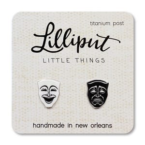 May include: A white card with black text that reads "Lilliput Little Things" and "handmade in new orleans". The card features two black and white enamel earrings with a comedy and tragedy mask design. The earrings are on a titanium post.