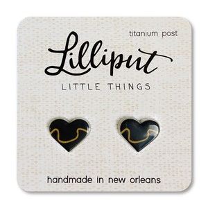 May include: A pair of black heart-shaped earrings with a gold line design. The earrings are on a white card with the text "Lilliput Little Things" and "titanium post" printed on it. The card also says "handmade in new orleans".