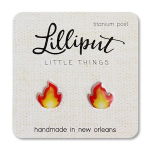 May include: A pair of flame-shaped earrings with a red and yellow design. The earrings are made of resin and have titanium posts. The earrings are handmade in New Orleans. "Lilliputit Little Things" is printed on the card.