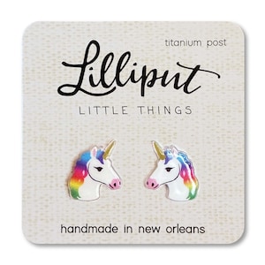 May include: A pair of rainbow unicorn stud earrings with titanium posts. The earrings are handmade in New Orleans and feature a white unicorn head with a rainbow mane and horn. The card reads "Lilliput Little Things".