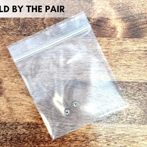 May include: A clear plastic bag containing two small, round, light-colored gemstones. The bag is sealed and sits on a wooden surface. Text at the top reads "SOLD BY THE PAIR".