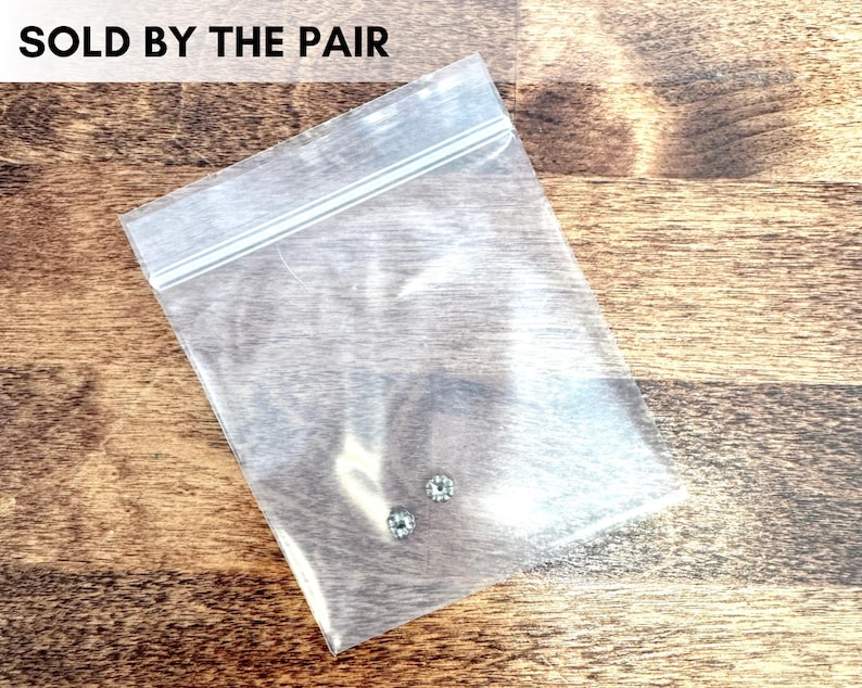 May include: A clear plastic bag containing two small, round, light-colored gemstones. The bag is sealed and sits on a wooden surface. Text at the top reads "SOLD BY THE PAIR".
