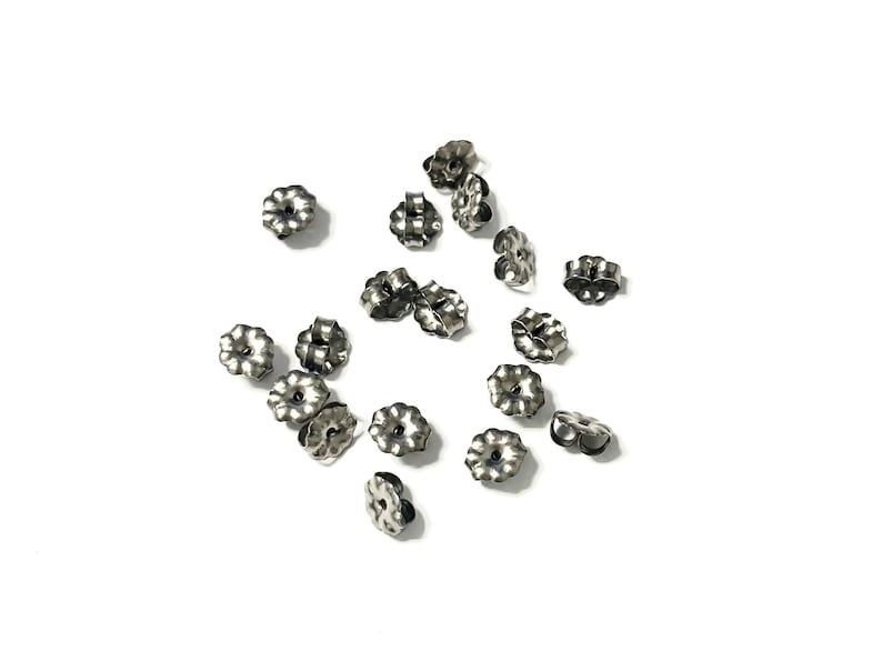 May include: A collection of silver-colored earring backs scattered on a white surface. The earring backs have a flower-like design with multiple petals and a small hole in the center. The backs are made of metal.