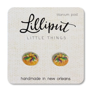 May include: A pair of earrings featuring a miniature, colorful, glazed donut with purple, green, and yellow icing. The earrings are handmade in New Orleans and have titanium posts. The card reads "Lilliputit Little Things" and "handmade in New Orleans".