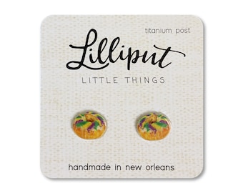 May include: A pair of earrings featuring a miniature, colourful, glazed donut with purple, green and yellow icing. The earrings are handmade in New Orleans and have titanium posts. The card reads "Lilliputit Little Things" and "handmade in New Orleans".