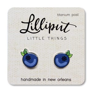May include: A pair of blueberry stud earrings on a white card. The earrings are blue with green leaves and a black center. The card reads "Lilliput Little Things" and "handmade in new orleans."