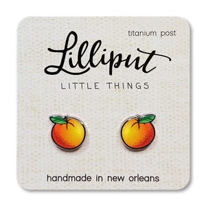 May include: A pair of orange fruit stud earrings with green leaves on a white display card featuring the text Lilliput Little Things and handmade in new orleans with a titanium post label. The earrings are round with a gradient yellow to red color.