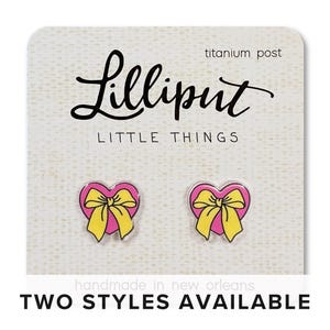 May include: A pair of pink heart-shaped earrings with yellow bows. The earrings are on a white card with the text "Lilliput Little Things" and "titanium post".