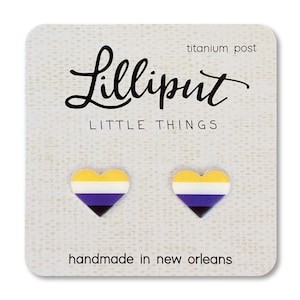 May include: Heart-shaped earrings featuring the non-binary pride flag colours: yellow, white, purple, and black. The card reads "Lilliput Little Things" and "handmade in new orleans". The earrings have titanium posts.