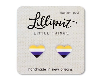 May include: Heart-shaped earrings with a non-binary pride flag design. The earrings are yellow, white, purple, and black. The card reads "Lilliput Little Things" and "handmade in new orleans". The earrings have titanium posts.