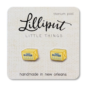 May include: A pair of butter-shaped earrings on a white card. The earrings are yellow with blue text that reads "Salted Butter." The card has the text "Lilliput Little Things" and "handmade in new orleans."