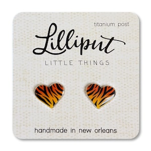 May include: A pair of heart-shaped earrings with a tiger stripe pattern in orange, black, and white. The earrings are handmade in New Orleans and have titanium posts. The card reads "Lilliput Little Things".