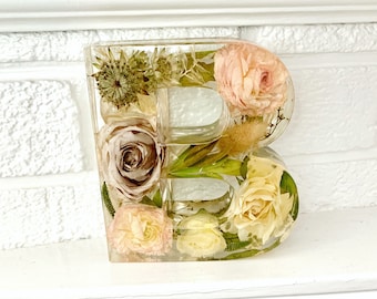 May include: A clear resin letter "B" filled with dried flowers in shades of pink, cream, and brown. The flowers are arranged within the letter's form, creating a decorative floral design. The letter is displayed on a white surface.