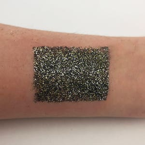 May include: A swatch of black and gold glitter applied to the skin. The glitter is densely packed, creating a shimmering, textured effect. The swatch is rectangular and appears to be a makeup product, likely eyeshadow or body glitter.