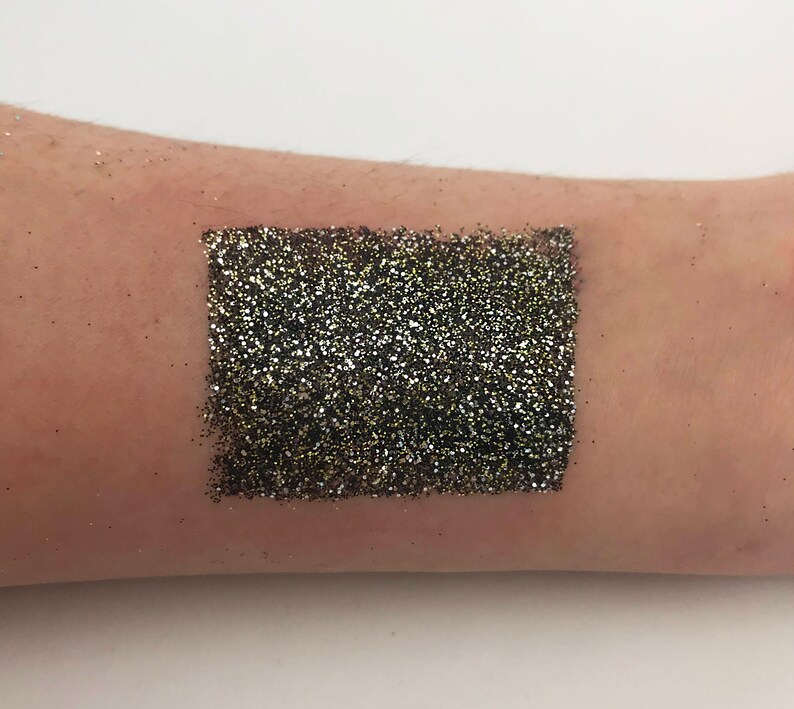May include: A swatch of black and gold glitter applied to the skin. The glitter is densely packed, creating a shimmering, textured effect. The swatch is rectangular and appears to be a makeup product, likely eyeshadow or body glitter.