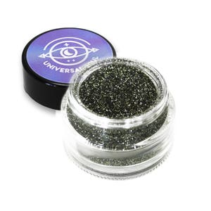 May include: A clear glass jar filled with dark green glitter, with the lid off to the side. The lid is black with a purple and blue label that reads "UNIVERSAL". The glitter sparkles and reflects light.