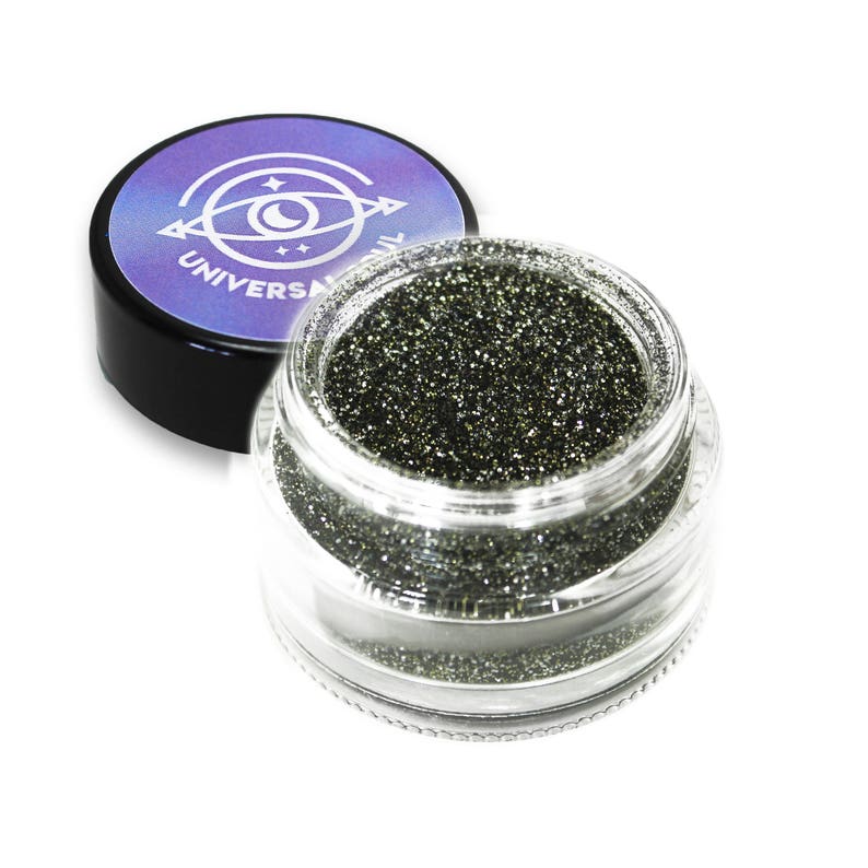 May include: A clear glass jar filled with dark green glitter, with the lid off to the side. The lid is black with a purple and blue label that reads "UNIVERSAL". The glitter sparkles and reflects light.