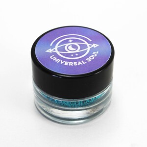 May include: A small, clear jar with a black lid, filled with sparkling teal glitter. The lid features a purple and blue label with a white eye design and the words "UNIVERSAL SOUL". The jar is set against a white background.