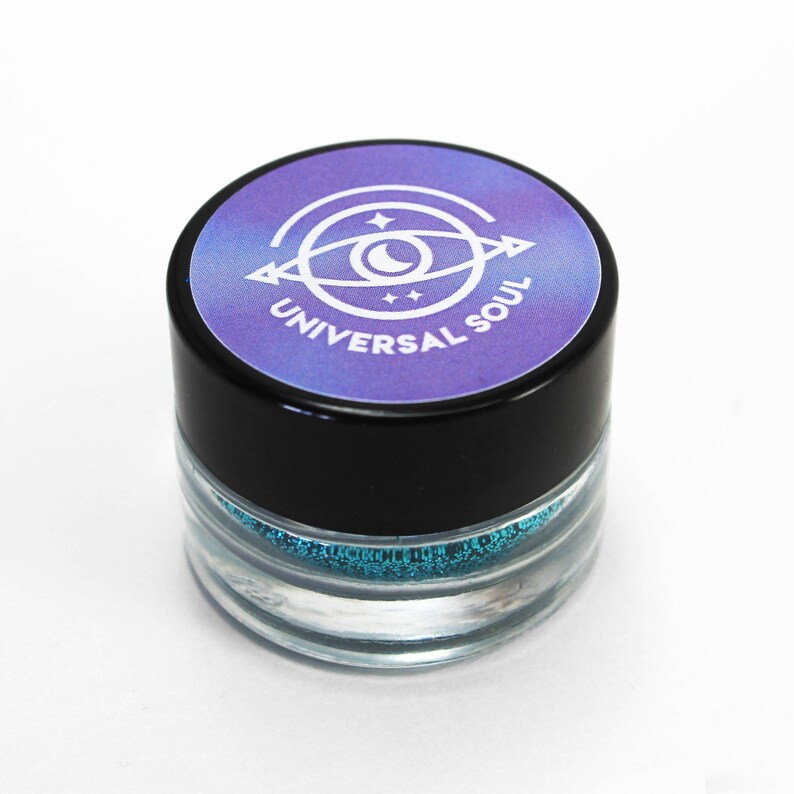 May include: A small, clear jar with a black lid, filled with sparkling teal glitter. The lid features a purple and blue label with a white eye design and the words "UNIVERSAL SOUL". The jar is set against a white background.