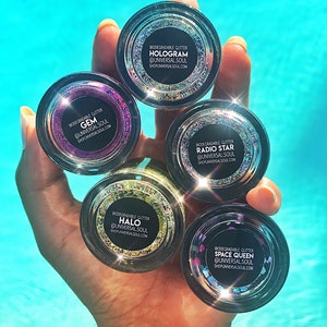 May include: A hand holds five small, clear jars of biodegradable glitter. Each jar has a black label with white text indicating the glitter type: Hologram, Gem, Radio Star, Halo, and Space Queen. The glitter colors vary, with a blue water background.