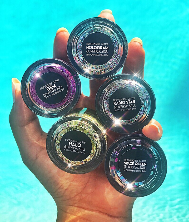 May include: A hand holds five small, clear jars of biodegradable glitter. Each jar has a black label with white text indicating the glitter type: Hologram, Gem, Radio Star, Halo, and Space Queen. The glitter colors vary, with a blue water background.