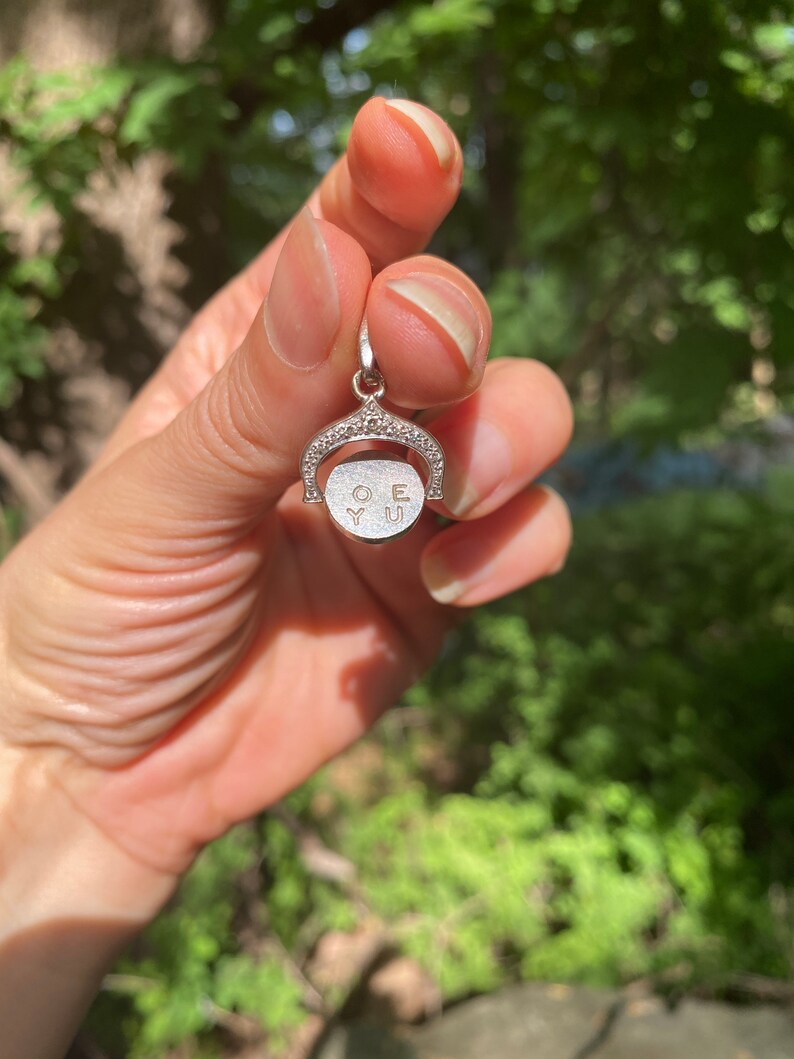 May include: A silver charm with a circle that rotates to reveal the words 'O.Y.U.' The charm is decorated with small, clear stones.