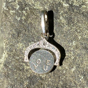 May include: A silver charm with a spinning disc that says 'LOVE YOU' in black lettering. The charm is decorated with small clear stones around the edge of the disc and the top of the charm.