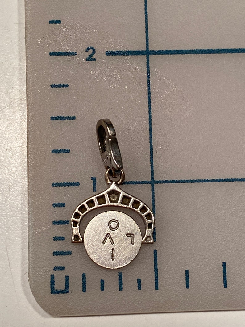 May include: A silver charm with a round disc featuring a circle, an arrow pointing up, and a number 1. The charm is attached to a silver loop.