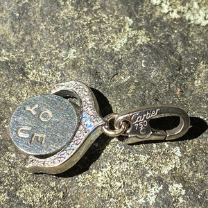 May include: A silver charm with a round disc that says 'XO YOU' in cursive. The charm is attached to a silver clasp that says 'Cartier 750'.