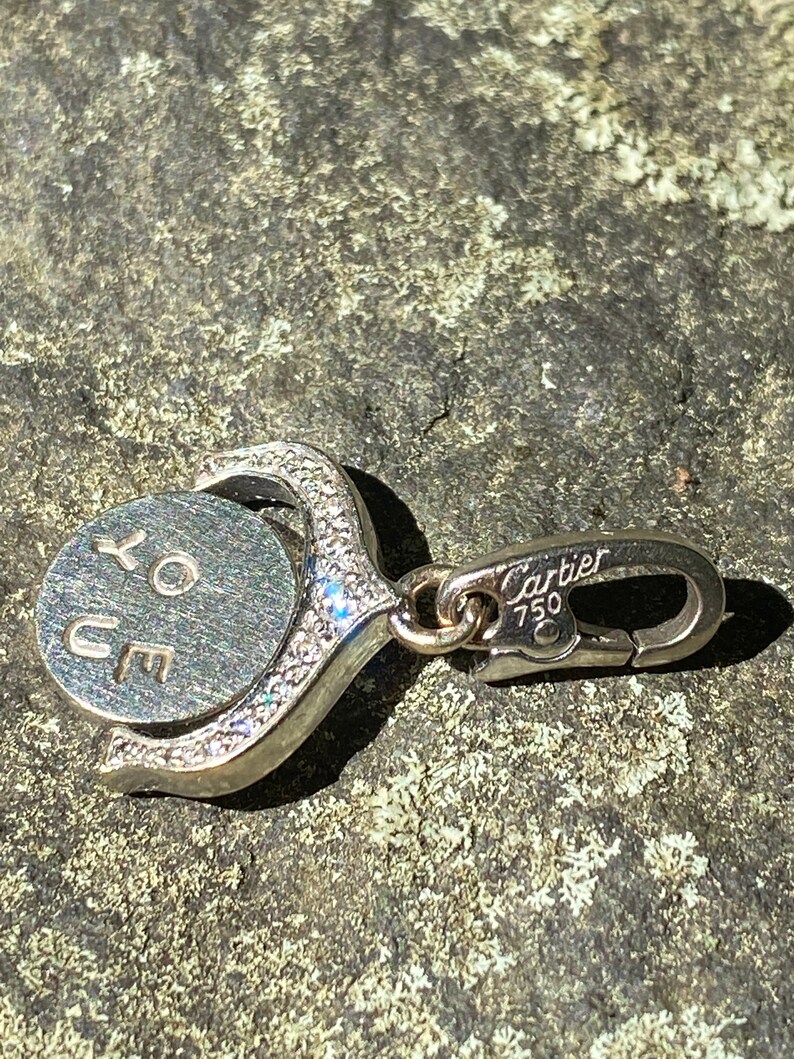 May include: A silver charm with a round disc that says 'XO YOU' in cursive. The charm is attached to a silver clasp that says 'Cartier 750'.