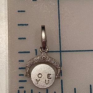 May include: A silver charm with a spinning disc that reads 'O.Y.U.E' on a white background. The charm is surrounded by small clear stones.
