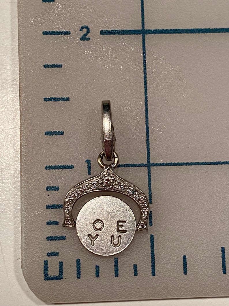 May include: A silver charm with a spinning disc that reads 'O.Y.U.E' on a white background. The charm is surrounded by small clear stones.