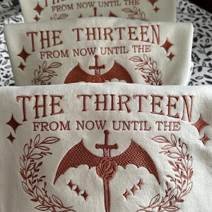 May include: Off-white sweatshirts with embroidered text and graphics. The text reads "THE THIRTEEN FROM NOW UNTIL THE DARKNESS". The design includes a sword, wings, and floral elements in a rust colour. The sweatshirts are stacked.