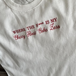 以下が含まれることがあります： 胸元に赤い刺繍で「WHERE THE F*** IS MY Cherry Float Coke Zero」と書かれた白い半袖Tシャツ。1行目は太字のサンセリフ体、2行目は筆記体のフォントで構成されています。