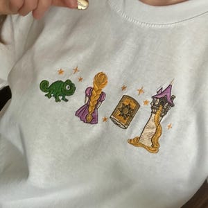 May include: White t-shirt with an embroidered design. The design includes a green chameleon, a blonde braid, a book, and a tower, in green, purple, gold, and orange. A gold necklace is visible at the top of the image.