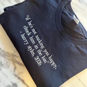 May include: A black crew neck t-shirt featuring white printed text that reads, "If he's not making you happy, chuck him in the bin" - harry styles, 2026. The garment is folded and displays a standard short-sleeve design with a visible neck label.