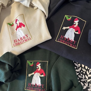 May include: Three crewneck sweatshirts in beige, black, and dark green, each featuring an embroidered rectangular patch. The patch depicts a figure in a red shirt and white trousers wearing a Santa hat, with holly and the text HARRY CHRISTMAS.