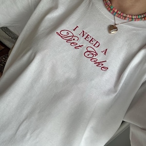 May include: A white short-sleeved t-shirt featuring the embroidered red text "I NEED A Diet Coke" on the chest. The shirt is paired with a multi-coloured beaded necklace and a gold-toned heart-shaped pendant on a thin chain.