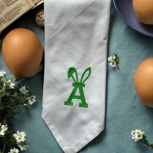 Può includere: Tovagliolo di stoffa bianco con una lettera "A" verde ricamata sormontata da orecchie di coniglio. Uova marroni, un uovo di quaglia e fiori bianchi circondano il tovagliolo su una superficie blu.