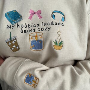 Puede incluir: Sudadera color crema con ilustraciones bordadas de un libro, un lazo, auriculares, café helado, una vela y una planta colgante. El texto "my hobbies include being cozy" está bordado debajo.