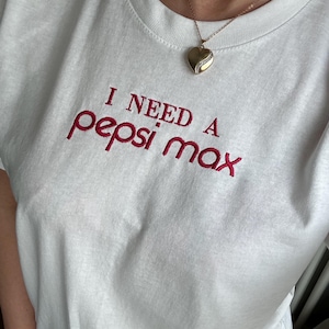 May include: A white t-shirt featuring the embroidered text "I NEED A pepsi max" in red thread. The shirt has a crew neckline and is paired with a gold-toned heart-shaped pendant necklace on a thin chain, resting against the fabric of the shirt.
