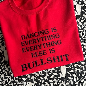 Könnte beinhalten: Ein leuchtend rotes Sweatshirt mit Rundhalsausschnitt und schwarzer Stickerei, die den Text DANCING IS EVERYTHING EVERYTHING EVERYTHING ELSE IS BULLSHIT zeigt. Das Kleidungsstück hat einen gerippten Kragen und lange Ärmel mit einem mittigen Schriftzug.