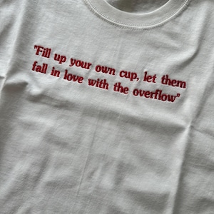 Może przedstawiać: Biały t-shirt z czerwonym haftowanym napisem: "Fill up your own cup, let them fall in love with the overflow". Koszulka posiada okrągły dekolt oraz metkę marki Fruit of the Loom widoczną wewnątrz przy kołnierzu.