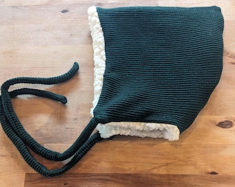 May include: A dark forest green bonnet featuring a textured knit exterior and a thick, cream-colored faux shearling lining. The headwear includes two long, matching dark green knitted ties attached to the sides for securing under the chin.